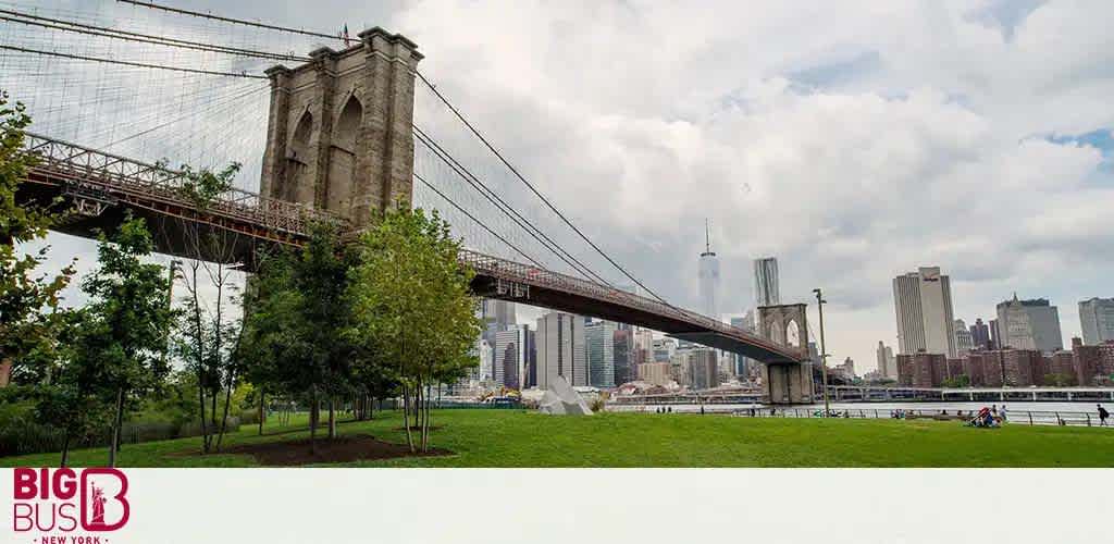 An overcast sky looms over the Brooklyn Bridge extending across the image with its distinctive stone towers and suspended cables. In the foreground, a green park with trees and visitors provides a relaxed setting against the bridge and the distant urban skyline of downtown Manhattan. The Big Bus New York logo sits prominently on the lower left.