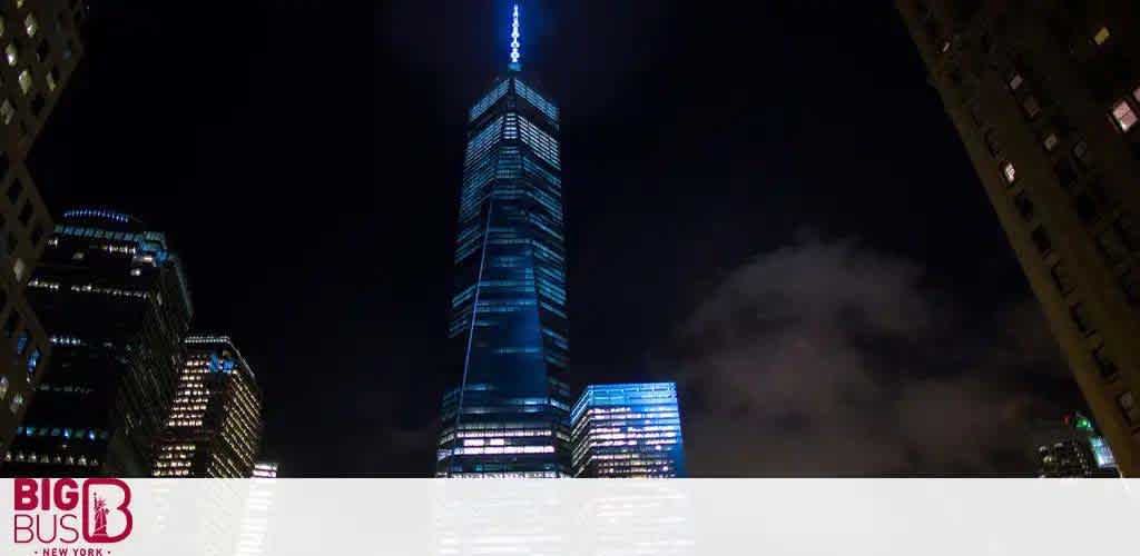 A nighttime view of a brightly lit skyscraper towering over surrounding buildings. The sky is dark with clouds faintly visible. Advertisements can be seen at the bottom of the image. The skyscraper stands out with its blue and white lights against the cityscape.