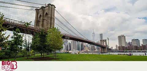 Image displays the Brooklyn Bridge spanning the East River, with the New York City skyline in the background. Waterfront park area with lush green grass is visible in the foreground, and the sky is partly cloudy. The bridge's intricate cable design and massive stone towers are prominent. A logo for Big Bus Tours is placed in the corner.