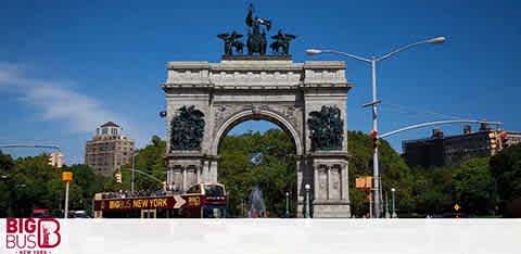 Image shows the Grand Army Plaza in New York with a large, ornate arch, topped by a sculpture group. In front, a red sightseeing bus is visible. Clear blue skies and trees are in the background with city buildings flanking the sides.