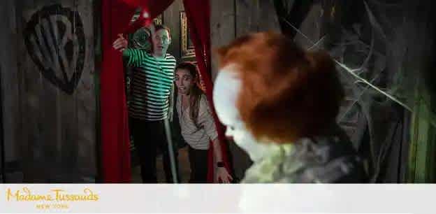 Image description: Two individuals express surprise and fear as they peek behind a red curtain where a figure with bright orange hair resembling a doll is seen from the back. The environment suggests a spooky or themed attraction, indicated by dim lighting and eerie decor. A logo on the bottom left suggests the setting is Madame Tussauds in New York.