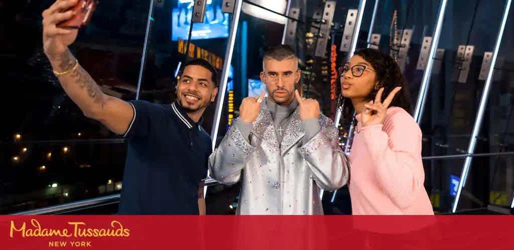 Image shows three individuals posing for a selfie at night. Two people on the sides are smiling and making hand gestures while the person in the middle, wearing a sparkly silver jacket, is giving a thumbs-down sign. In the background, city lights are visible. The Madame Tussauds New York logo is in the bottom left corner.