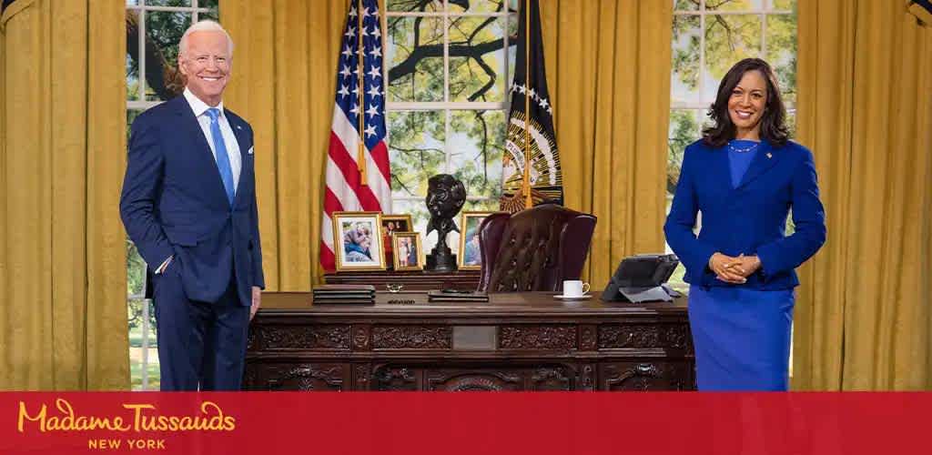 Image displays life-sized wax figures in an Oval Office setting. A man stands on the left, wearing a blue suit, white shirt, and tie, smiling at the camera. A woman on the right, also in a blue suit with her hands clasped, beams at the viewer. American flag and desk with photos in the background. Madame Tussauds New York logo at the bottom.