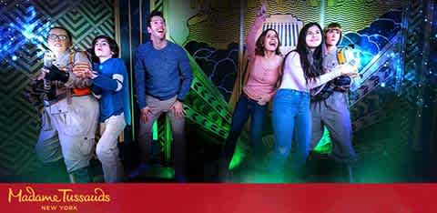 This image features four excited individuals posing together at Madame Tussauds New York against a colorful backdrop with dynamic lighting effects reminiscent of a sci-fi theme. They are acting surprised and thrilled, as if engaging in an interactive exhibit. A mix of casual attire suggests a relaxed, fun atmosphere. The Madame Tussauds logo is visible in the bottom corner.