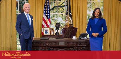 Image of two figures resembling public figures standing in an oval office setting. One figure is on the left in a blue suit, standing with hands clasped, the other on the right in a blue dress, smiling towards the camera. The room has curtains, a desk with a chair, flags, and the logo of Madame Tussauds New York in the corner.