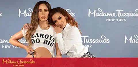 Image shows two individuals posing side by side at Madame Tussauds New York, against a backdrop with the attraction's logo. The person on the left wears a cropped top with the phrase 'GAROTA DO RIO' and is striking a poised stance, while the one on the right, dressed in a chic ensemble, leans in with a smile. Both exude a playful yet stylish vibe.