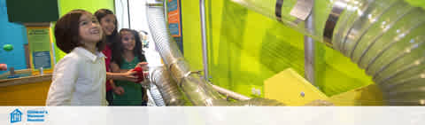 Image description: Three children are engaged in an educational activity at a science center. They are focusing on a large, coiled metallic tube, with curious expressions. Bright green and yellow walls with interactive displays add to the lively learning environment.