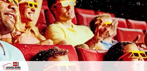 An excited audience wearing yellow 3D glasses at a LEGOLAND Discovery Center theater. People of various ages are smiling and reaching out, as if interacting with the on-screen action. The room has a warm glow and each seat appears comfortable and spacious. The LEGOLAND logo is visible in the bottom right corner, indicating the branded entertainment experience.