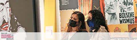 Two individuals, one in a black mask and the other in a blue mask, are standing in a gallery with historical posters on the wall. The text indicates the location is the Dallas Holocaust and Human Rights Museum. The ambiance suggests an educational exhibit.