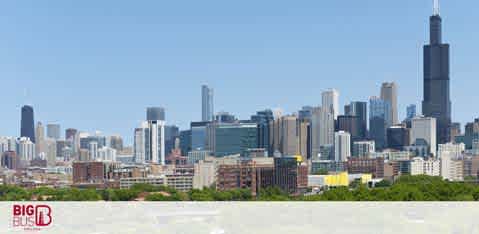 The image depicts a clear view of a city skyline on a bright day. Skyscrapers of various heights form a dense urban landscape, with the tallest building dominating the center. The foreground is blurred, while the sky is a clear blue. The bottom edge features a red logo with white lettering.