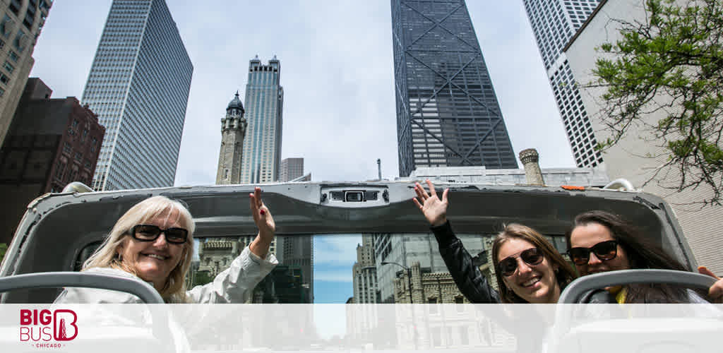 Three joyful tourists are enjoying a sightseeing tour on a Big Bus in Chicago. Two younger women and an older woman wave and smile brightly, exhibiting the fun of urban exploring. The sunny weather and clear skies complement the towering skyscrapers surrounding them, reflecting the excitement of the city tour.