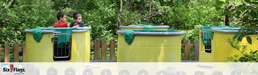 Image displays a children's theme park ride featuring bright yellow, barrel-shaped cars on a track, partially concealed by greenery in the background. Two children are visible looking out from the lead car. The Six Flags logo is apparent at the bottom of the image, indicating the location is a Six Flags amusement park.