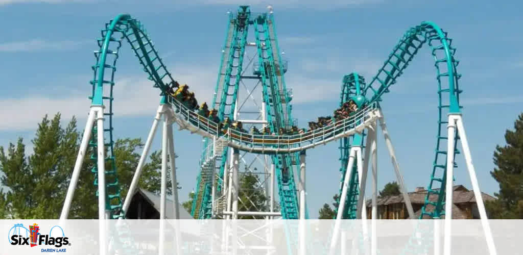Image shows a towering roller coaster with a series of inverted loops and drops in turquoise and white colors. The coaster is filled with riders and set against a clear sky, surrounded by trees. The Six Flags logo is prominently displayed at the bottom.