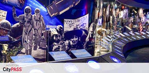 This image displays an exhibition space related to space exploration. On the left are two astronaut figures in spacesuits next to a photograph of an astronaut with a flag. On the right, informational panels are spread out with a projector screen displaying the phrase 'Invasion of the Space Invaders' and a person's image. 'Houston CityPASS' is highlighted at the bottom left.