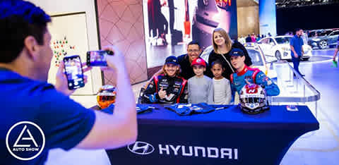 Image Description: This photograph captures a lively moment at an auto show, specifically at a Hyundai promotional booth. In the foreground, we see the back of a person taking a picture with a smartphone, framing a group of people. The focus of the image is on the group at the center, consisting of two individuals dressed in racing attire, complete with branded caps and seated behind a table. Standing between them are three young attendees, smiling for the photo, accompanied by a woman positioned just behind with a joyous expression also taking part in the photo opportunity. In the background, the well-lit exhibition hall reveals the outlines of various cars and auto show attendees, with the Hyundai logo prominently displayed on the front of the table and on a sign in the mid-background. The Auto Show logo is visible in the bottom left of the image.

Incorporate this exciting experience into your next outing with FunEx.com, where you can always count on huge savings and the lowest prices on tickets to events like these.