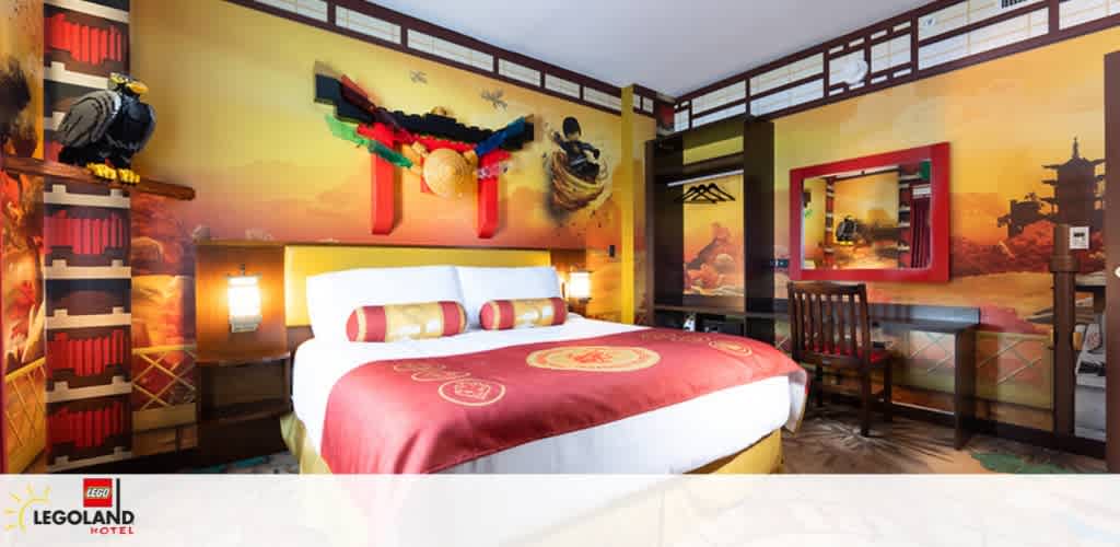 Image shows a vibrant Legoland Hotel room with a ninja theme. The walls feature colorful murals with Ninjago characters, and the room includes a plush double bed with themed beddings. A LEGO-style weapon hangs above a bunk bed, and there's a desk area with a chair. A large window provides natural light, completing the playful atmosphere.