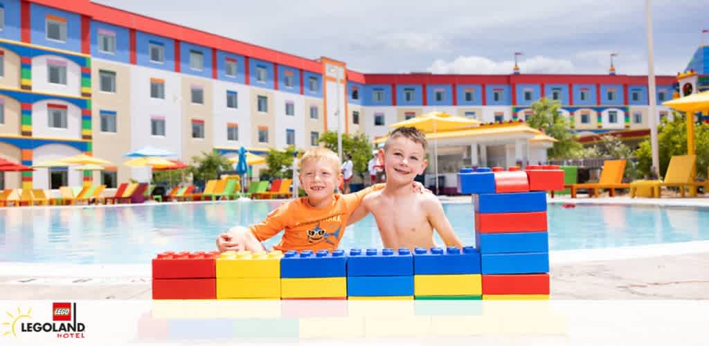 Two children play with large, colorful building blocks by a pool in front of the vibrant, multicolored Legoland Hotel. The sky is partly cloudy, and the pool area is adorned with bright umbrellas and loungers, reflecting a cheerful, family-friendly atmosphere.