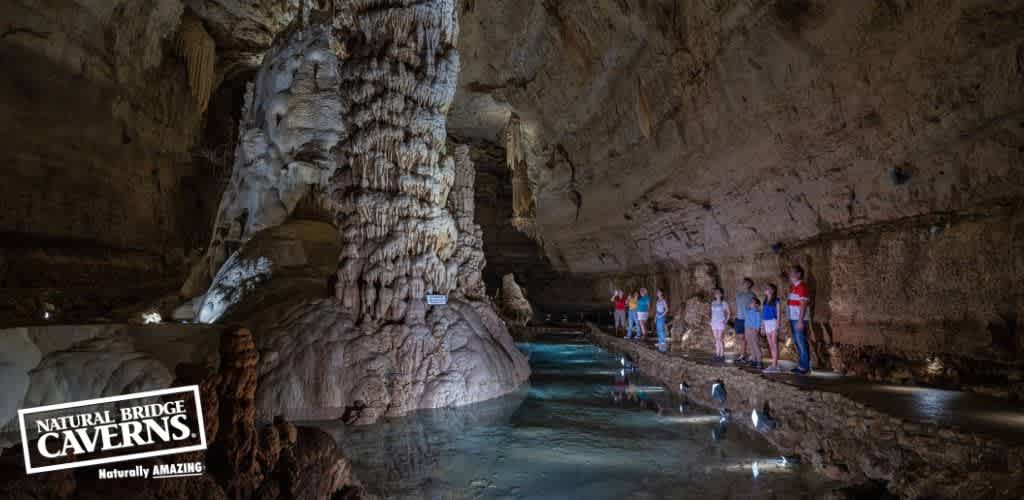 An expansive underground cavern with a group of visitors standing on a walkway. Majestic rock formations and clear, still water reflect the dim, artificial lighting, highlighting the natural beauty of the cave. The location is identified as Natural Bridge Caverns.