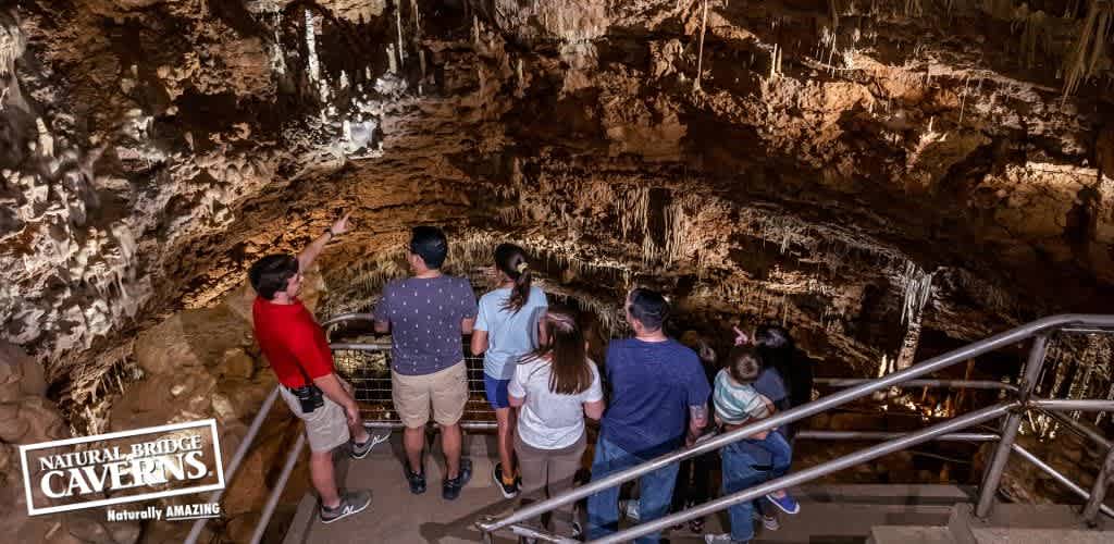 A group of visitors is exploring the textured interiors of Natural Bridge Caverns. They stand on a metal walkway, attentively observing the intricate formations. The cavern around them showcases an array of stalactites and stalagmites. The image includes the logo  Natural Bridge Caverns - Naturally AMAZING  in the foreground.