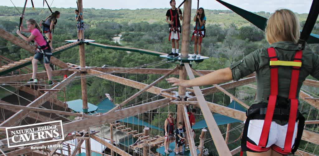 Visitors are securely harnessed while navigating an outdoor high-ropes adventure course. Wooden platforms and beams are suspended against a backdrop of lush greenery. The Natural Bridge Caverns logo with  Naturally AMAZING  slogan is visible in the corner.