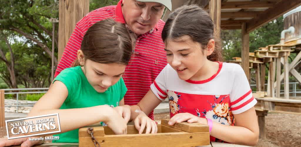 A man in a red shirt and two girls, one in green and the other in a white and red striped shirt, are looking into a wooden box at Natural Bridge Caverns, surrounded by lush trees and wooden structures. They appear engaged and focused on their activity.