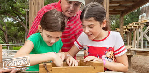 An adult and two children engage in an educational outdoor activity. They appear focused on items in a wooden container, possibly gemstone panning. Green foliage and a rustic structure set a natural background, with text referencing Natural Bridge Caverns.