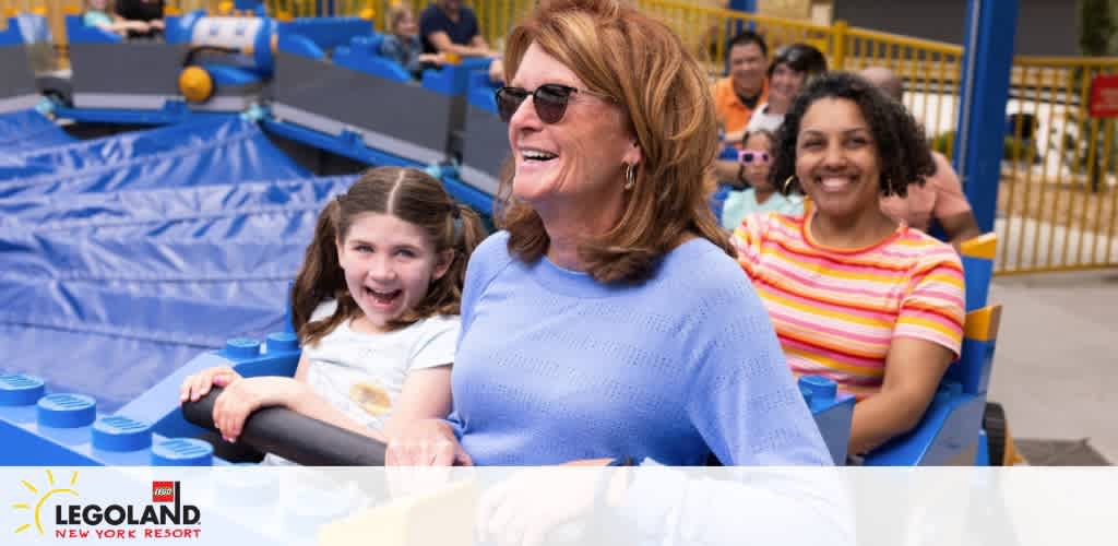 Visitors enjoying a ride at LEGOLAND New York Resort. A happy child and two women, one older with glasses and a younger one with curly hair, are smiling. They are seated in a blue attraction resembling LEGO bricks. Background shows other guests and bright yellow railings.