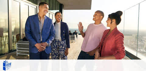 This image features four people on a balcony with a clear sky backdrop. Two men and two women, dressed in business casual attire, are engaged in a cheerful conversation. The man on the left wears a blue suit and smiles while talking to a woman in a red jacket who is facing him. Facing the camera is a woman in a white blouse and patterned skirt, both laughing with the man in the center wearing a pink shirt. The environment suggests a relaxed, professional setting.