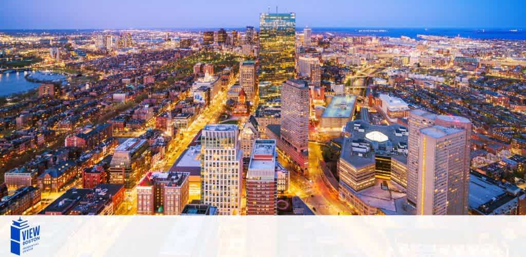 Panoramic view of a city skyline at dusk. Bright streetlights line the bustling roads while the buildings are illuminated from within. The horizon shows a twilight sky transitioning from blue to dark with distant lights visible along the waterfront. A logo reads VIEW Boston at the bottom left.