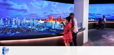 The image features an observation deck with people overlooking a panoramic cityscape at dusk. The city lights create a vibrant contrast against the twilight sky. A man and woman stand close to the railing on the left, while another person is seen at a distance on the right. The space is illuminated by the large screen displaying the city view and ambient lighting along the deck. A logo with 'THE VIEW' is visible in the bottom left corner.