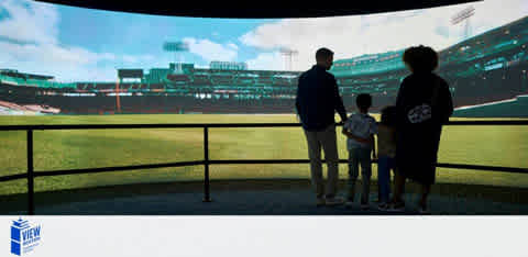 Image shows a silhouette of a family of four standing in front of a large panoramic window overlooking a baseball stadium. The stadium is well-lit by daylight with visible seating tiers and a vibrant green field. The family seems to be enjoying the view on a sunny day.