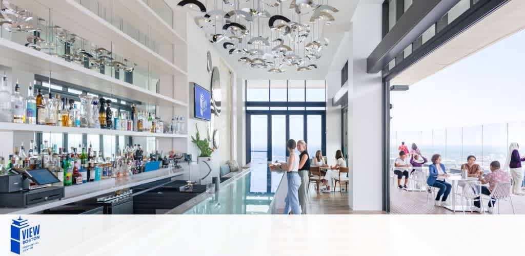 An elegant bar with a white marble countertop is stocked with various bottles. Modern chandeliers hang above. An adjacent area with floor-to-ceiling windows overlooks the ocean and includes patrons seated at tables, engaging in conversation.