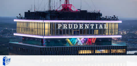 The image displays the illuminated upper section of a skyscraper at dusk. The building is prominently labeled with the word 'PRUDENTIAL' in large letters. Below, colorful neon signs light up the facade. The surrounding view suggests the building is located within a large cityscape.