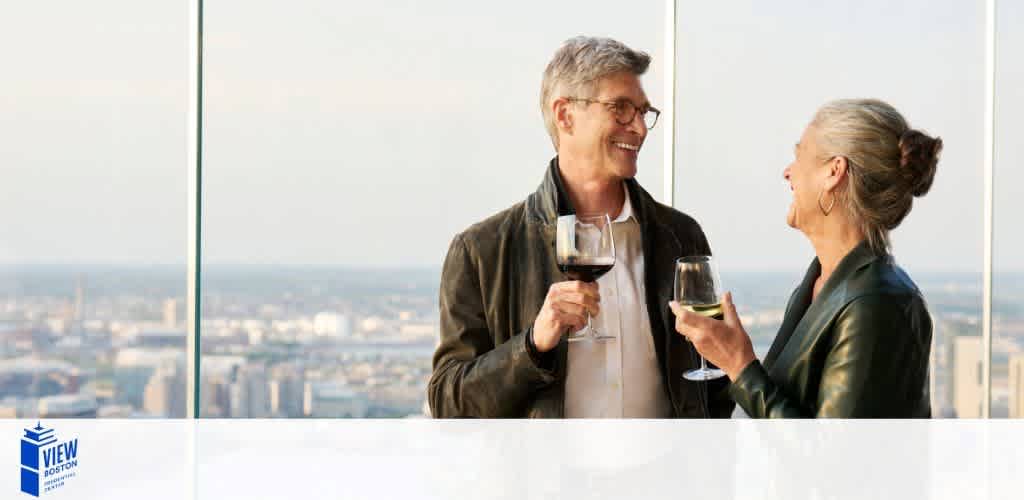 Image shows a man and woman in casual clothing, cheerfully toasting with wine glasses against a backdrop of a city view through floor-to-ceiling windows. The setting suggests a high-rise building with a panoramic view. The logo for 'VIEW Boston' is visible in the corner.