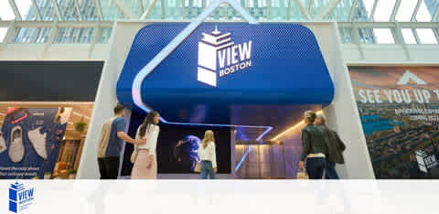 The image shows a group of people approaching a large promotional booth for  VIEW Boston . The booth's design is reminiscent of an oversized tablet in landscape orientation, with a deep blue upper half featuring the logo and a lighter blue arrow-shaped motif pointing towards the entrance. The environment suggests an indoor venue with a glass ceiling.
