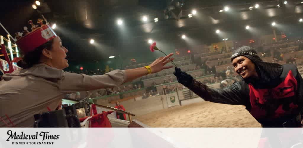 Image description: This image captures a heartwarming moment at a Medieval Times Dinner & Tournament event. On the left, a woman dressed in Medieval-style servant attire, complete with a headdress, extends her arm to present a single red rose. On the right, a man adorned in a knight's costume, featuring chainmail and a cowl, reaches out joyfully to accept the rose. His expression is one of delight and appreciation. They are both illuminated by warm lighting that emphasizes the festive atmosphere. In the background, an arena filled with spectators seated at tiers is faintly visible, contributing to the immersive medieval ambiance. The Medieval Times logo is visible in the lower-left corner, indicating the theme of the entertainment.

For an epic journey back to the days of valor and chivalry, get your tickets at FunEx.com, where you'll find the lowest prices and biggest savings for a memorable medieval experience.