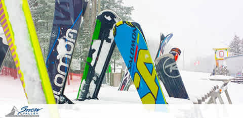 Image description: This is a photograph depicting a collection of skis and snowboards vertically inserted into the snow, with the tips pointing upwards. They are clearly arranged for storage while not in use, possibly by individuals taking a break from skiing or snowboarding. The equipment features a variety of colorful designs, including bright yellows, blues, greens, and blacks, with some displaying prominent branding. In the background, a winter scene is visible with lightly falling snow, and the setting appears to be a ski resort given the presence of signage that includes the Snow Valley logo. There are additional elements in the background that suggest this location is dedicated to winter sports, such as what may be a lift line area and infrastructure related to ski slopes. 

At FunEx.com, experience exhilarating wintertime adventures at the lowest prices available—we're your go-to source for significant savings on tickets to the most exciting snowy escapes!