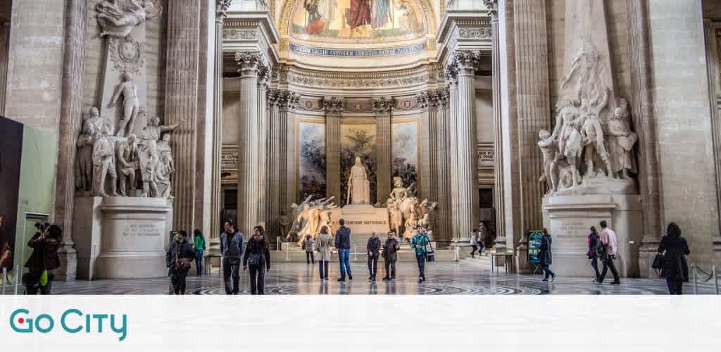 Visitors explore a grand neoclassical hall with soaring columns and intricate sculptures. Sunlight spills through the high windows onto marble floors. In the center, an imposing statue draws the eye amid the spacious, ornate architecture. The Go City logo appears at the bottom left.
