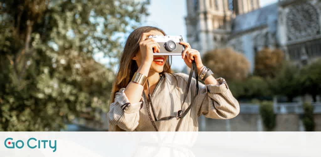 A joyful tourist takes a photo with a silver camera. She's outdoors and smiling behind the camera, near a cathedral along with trees in the background. The logo 'Go City' is in the corner, suggesting travel and sightseeing services.