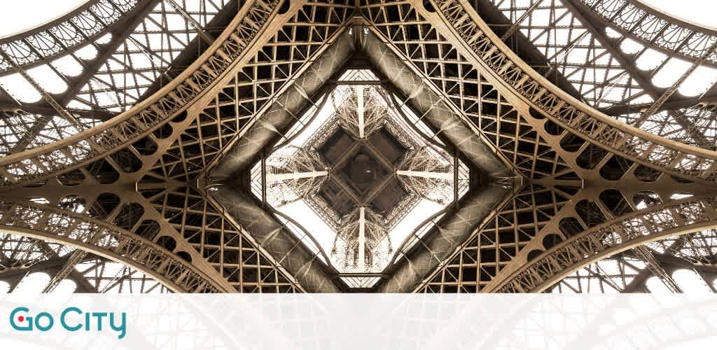 Symmetrical view from the base of the Eiffel Tower looking upwards, showcasing the intricate iron lattice work, with the Go City logo at the bottom center. The sky peeks through the structure's patterns.
