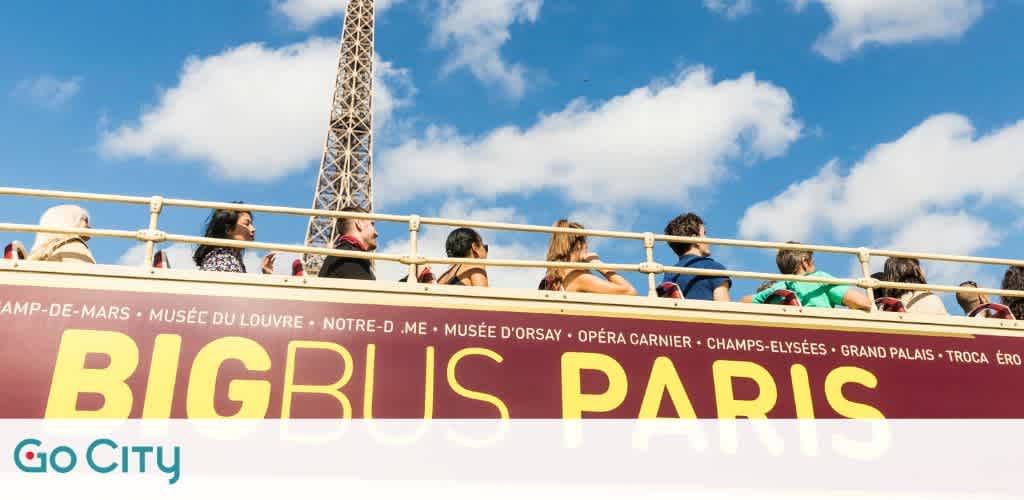 Image shows a clear day with people on the upper deck of a Big Bus tour bus. Large, bold text 'BIG BUS PARIS' is visible on a maroon background with the list of Parisian landmarks. Part of the Eiffel Tower is seen in the top left, suggesting the bus is near the famous structure. The Go City logo is at the bottom.