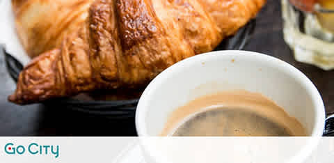 A fresh, flaky croissant accompanied by a cup of coffee with a light layer of crema on top. Both items are set on a table suggestive of a morning breakfast. The Go City logo appears in the corner, signaling a city lifestyle theme.