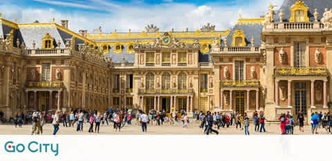 Image shows the exterior of the opulent Palace of Versailles under a bright blue sky with fluffy clouds. Visitors gather in the spacious courtyard, some walking while others take photos. The palace's architecture boasts ornate golden statues and intricate designs. The Go City logo is at the bottom right.