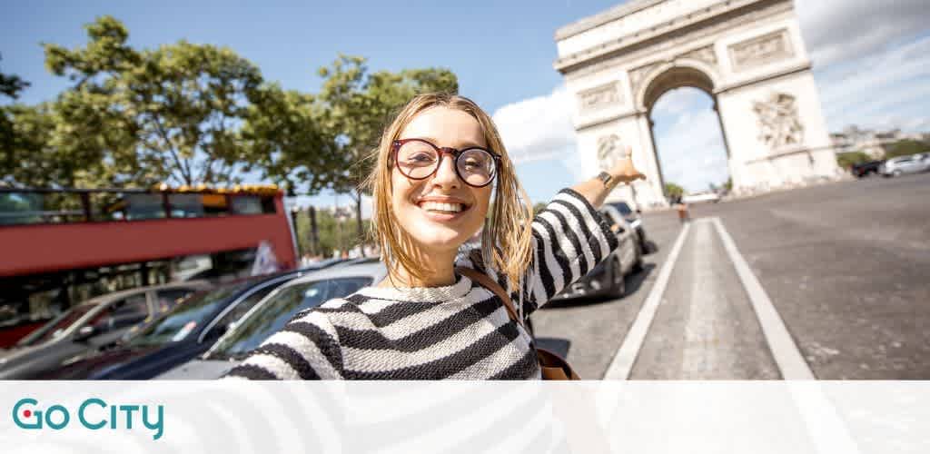 A woman with glasses takes a selfie in front of a famous arch in a city, smiling and pointing towards it. She wears a striped shirt, with cars and a red double-decker bus visible in the background. The sky is clear and sunny, and trees are lining the street.