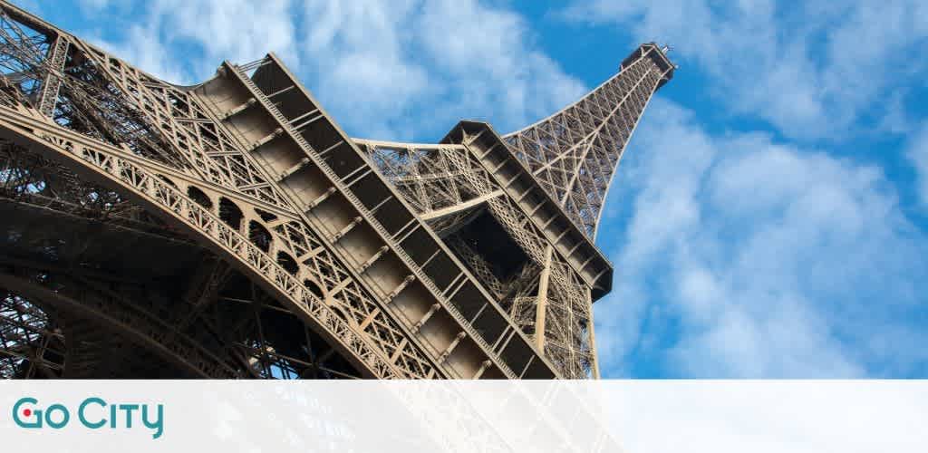 View of the Eiffel Tower from below set against a bright blue sky with scattered white clouds. The intricate iron lattice of the tower is prominently visible, showcasing its architectural detail. The image includes a Go City logo in the corner, suggesting city tourism or travel.