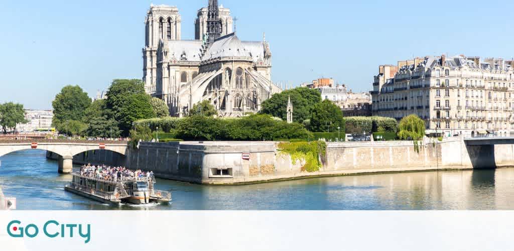A scenic view of a historic European cityscape featuring a grand cathedral with two towers. The river in the foreground has a boat filled with passengers enjoying a sunny day. Lush green trees and classic buildings line the riverbank, creating a picturesque urban scene.