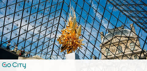 A golden, spiky sculpture is displayed inside a large, glass-paneled structure with a triangular roof. The sky is clear and blue, enhancing the dramatic architecture. Part of a historic stone building is visible in the background. The Go City logo is in the lower left corner.