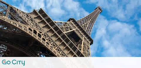 View of the Eiffel Tower from below against a blue sky with scattered clouds. The iconic iron lattice structure is angled dramatically upward, showcasing its intricate design. Sky provides a serene backdrop, enhancing the tower's grandeur.