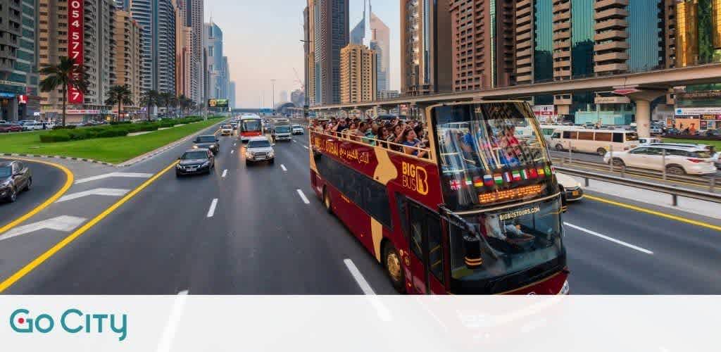 An open-top red tour bus filled with passengers travels on a city road flanked by modern high-rise buildings. Vehicles move in the contiguous lanes under a clear sky at dusk. The image conveys the experience of urban sightseeing. The logo Go City is present in the corner.