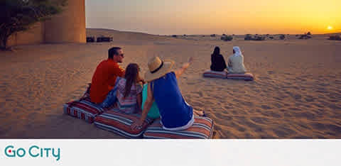 This image shows a serene desert landscape at sunset. Two pairs of people are seated on blankets enjoying the view. The foreground features a man in orange and a woman in blue, gazing towards the horizon. In the background, two individuals are observing the setting sun. The sky fades from warm amber to soft blue above the sandy expanse. The 'Go City' logo is at the bottom.